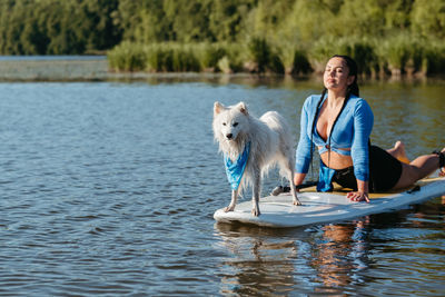 Snowy japanese spitz dog standing on sup board, woman doing stretching while paddleboarding