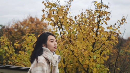 Young woman looking away while standing against trees during autumn