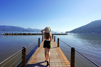 Rear view of woman standing by lake against clear blue sky