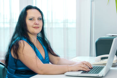 Brunette woman at home office working on laptop while looking at camera. horizontal photo