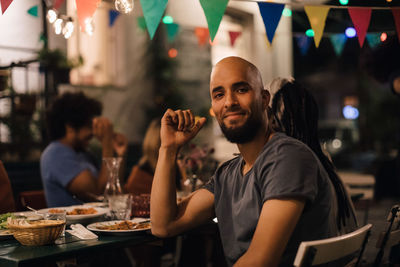 Portrait of smiling young man with shaved head sitting at table during dinner party in backyard