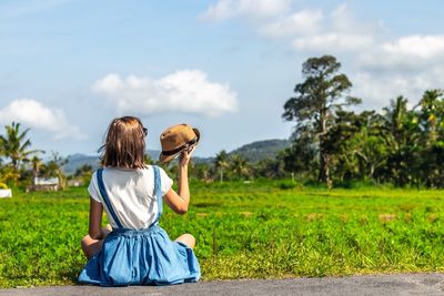 Rear view of woman holding hat while sitting at farm against sky