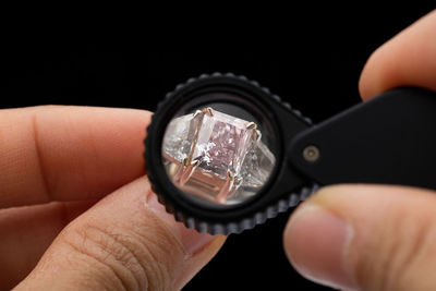 Cropped hands holding diamond ring with loupe over black background