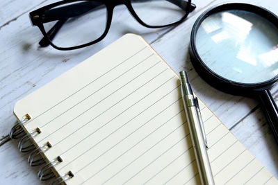 High angle view of note pad with pen by eyeglasses and magnifying glass on table