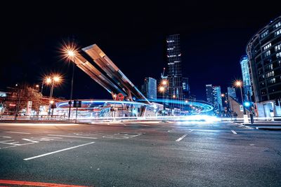 Light trails on city street by buildings against sky at night