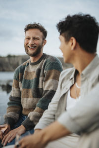 Happy young man enjoying with male friend while sitting near lake