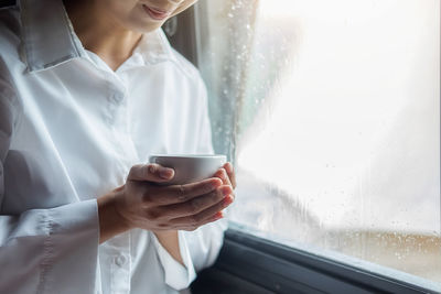 Midsection of woman having coffee while sitting by wet window at home