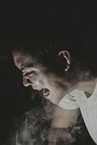 Side view of angry young man emitting smoke against black background