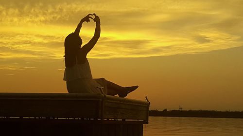 Rear view of woman making heart shape by sea on pier during sunset