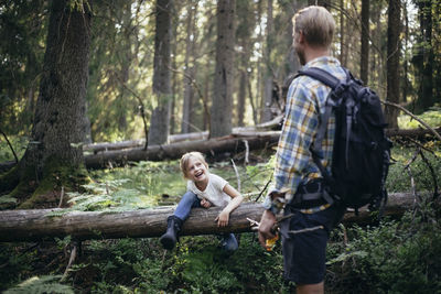 Smiling daughter climbing over fallen tree while looking at father in forest