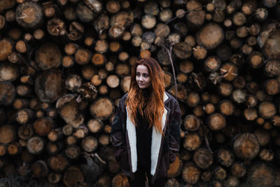 Young red haired female in warm jacket standing against stacked wooden logs in autumn day in countryside