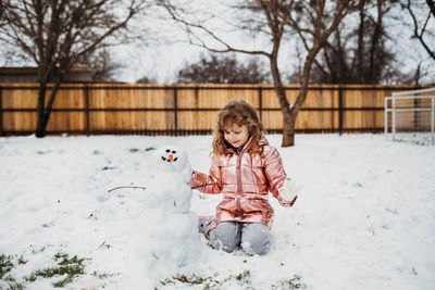 Young girl sitting in snow happily looking at snowman in winter
