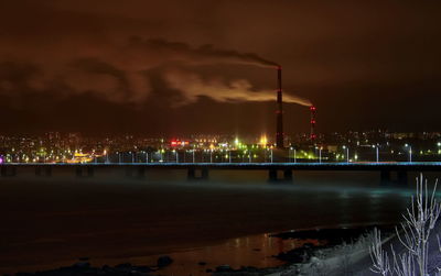 Illuminated factory by river against sky at night