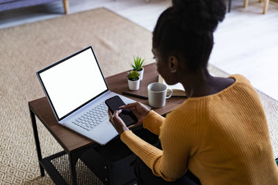 Afro woman using smart phone while sitting with laptop at home