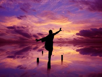 Rear view of silhouette person balancing on wooden post at lake against dramatic sky