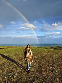 Full length of young woman standing on field against rainbow in sky