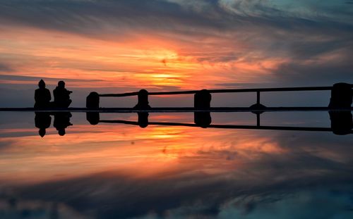 Symmetric view of silhouette friends sitting at lakeshore against cloudy sky during sunset