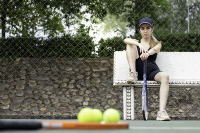 Frontal portrait of girl sitting next to tennis court waiting with racket out of focus in foreground