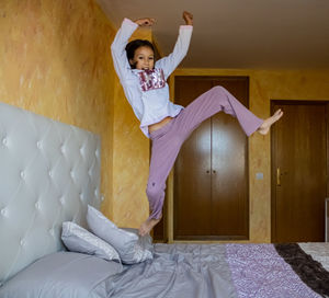 Portrait of girl jumping on bed against wall at home