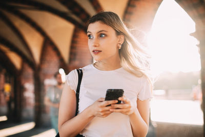 Young woman looking away while holding smart phone on footpath in city