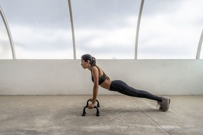 Side view of fit ethnic sportswoman in active wear exercising on push up bars while looking forward