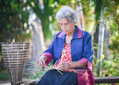 Woman making wicker baskets while sitting on bench against trees