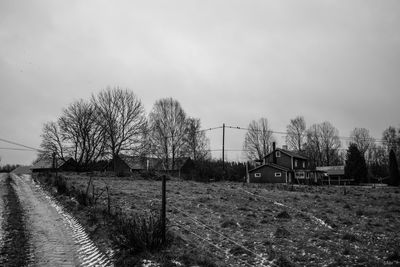 Bare trees on field by houses against sky