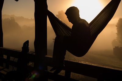 Man sitting on hammock against sky during sunset