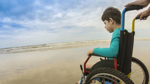 Side view of boy sitting in wheelchair at beach against sky