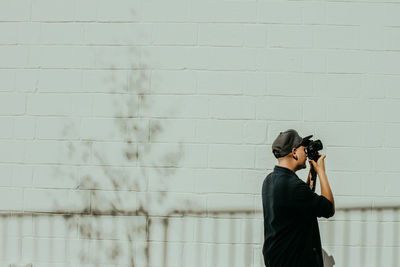 Side view of man photographing with camera against wall during sunny day