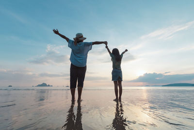 Rear view of mother and daughter holding hands while standing at beach against sky