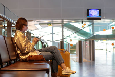 Side view of woman wearing mask using phone while sitting on chair at airport
