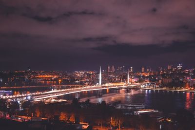 Illuminated bridge over river by buildings against sky at night