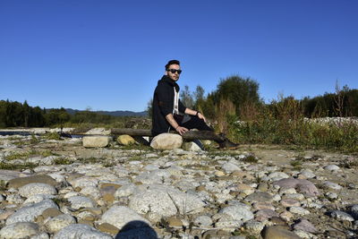 Portrait of young man sitting on rock against sky