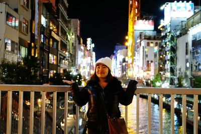 Portrait of happy woman gesturing while standing on bridge against illuminated buildings