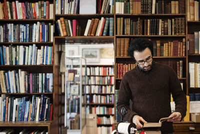 Librarian reading book while standing against shelves
