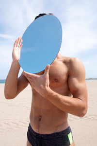 Shirtless man holding mirror while standing at beach against sky