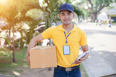 Portrait of smiling delivery person with box and clipboard standing on footpath