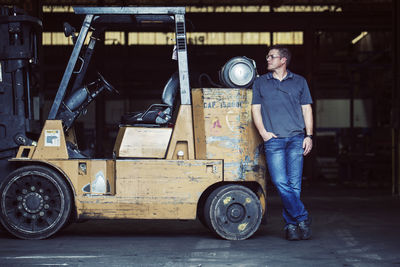 Full length of worker standing by forklift in industry