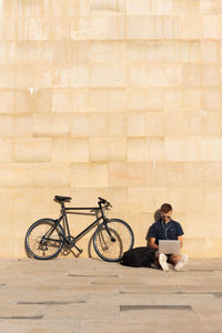 Young entrepreneur working outdoors on his laptop with his bicycle leaning on concrete wall