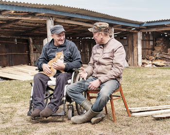 Happy senior man in wheelchair in yard of his house smiling holding cute cat, talking to his friend.