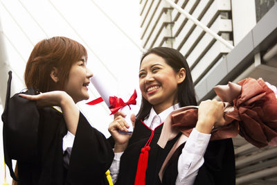 Cheerful female students wearing graduation gowns against building