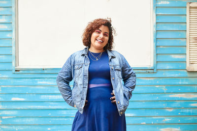 Pump cheerful female standing on street looking at camera on wooden blue cabin