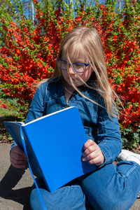 Young school age girl reading from blue textbook in park against bright red flowers background