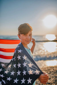 Portrait of boy holding american flag while standing at beach during sunset