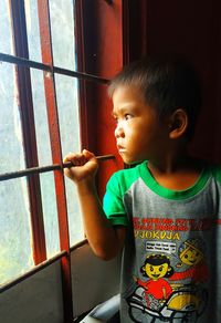 Cute boy looking away while standing against window at home