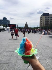 Cropped hand having flavored ice in city against sky