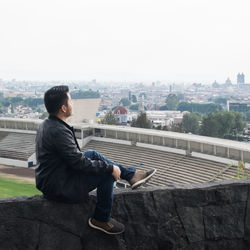 Side view of man sitting on retaining wall against cityscape