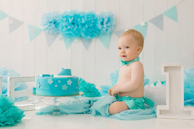 A one-year-old boy in blue shorts and a bow tie celebrates his birthday next to a cake on a stand 