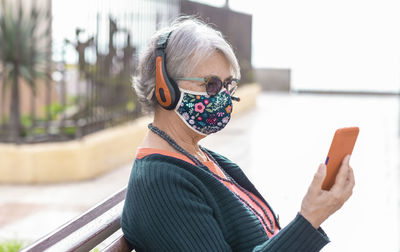 Close-up of senior woman wearing mask listening music while sitting outdoors